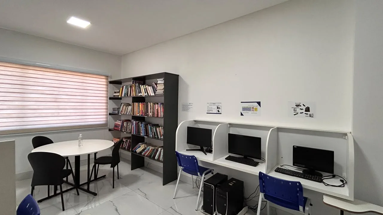 Sala com estante de livros, mesa de estudos e baias de computadores do laboratorio de informatica