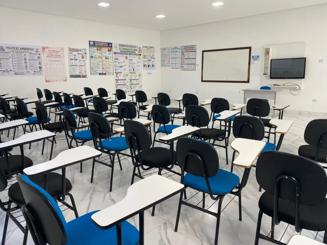 Sala de aula ampla com carteiras universitarias, quadro branco, televisao e paineis de farmacologia